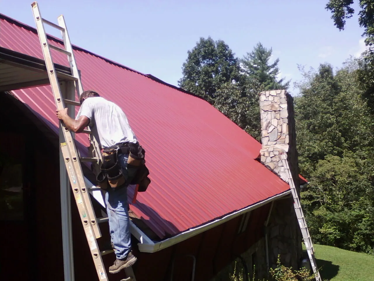 Roofer on a ladder beside a red metal roof and stone chimney — Soffit & Fascia Repair by Roofing Brothers in Poughquag